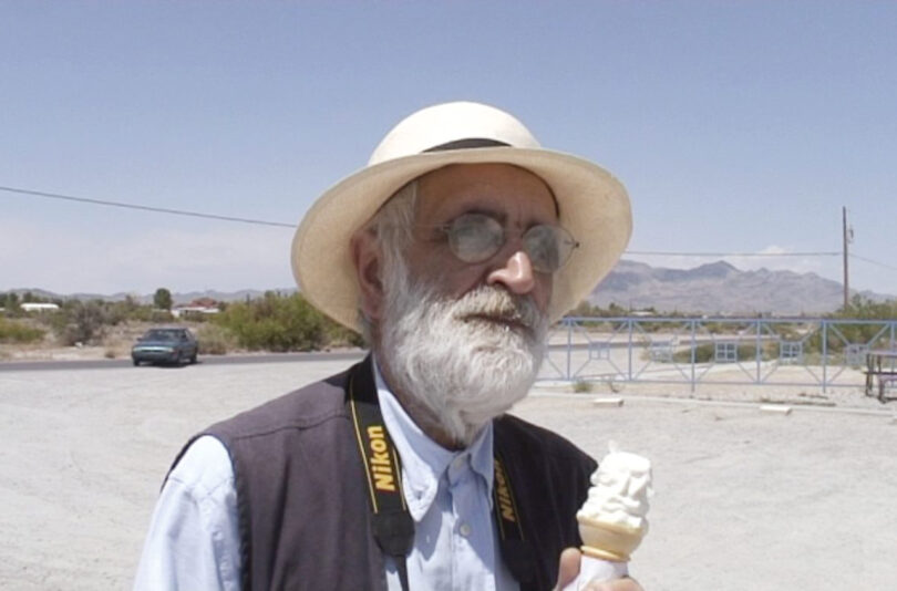 An elderly man wearing a white hat and glasses, holding an ice cream cone, stands in a barren parking area on a sunny day.