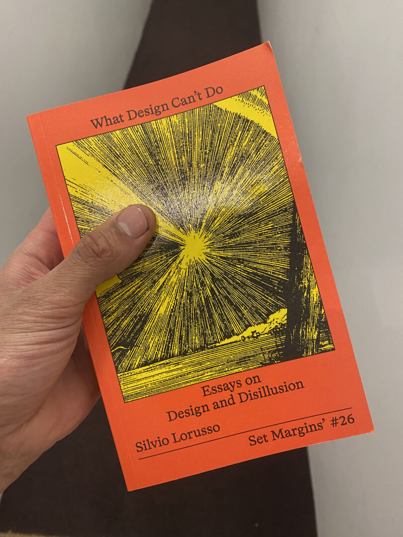 A hand holding a book titled "What Design Can't Do", featuring a bright yellow burst graphic on an orange cover.