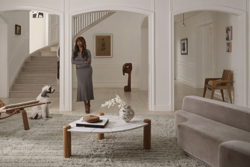 A woman in a gray dress stands in a living room with a dog.