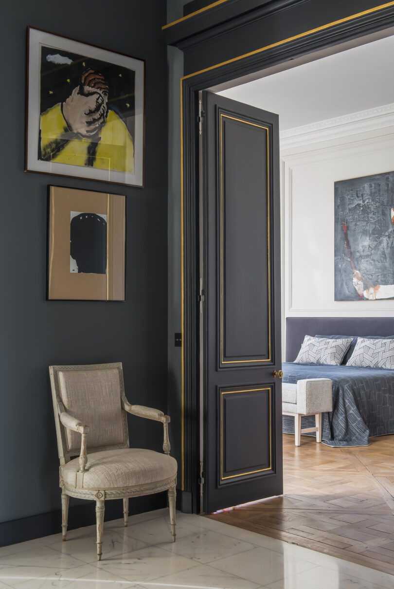 A room with dark walls and a vintage chair, featuring modern artwork and a peek into an adjoining room with a bed.