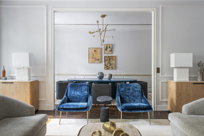Living space featuring plush blue chairs, gold and white lighting fixture overhead, flanked by large wooden cabinets and accented with modern decor.