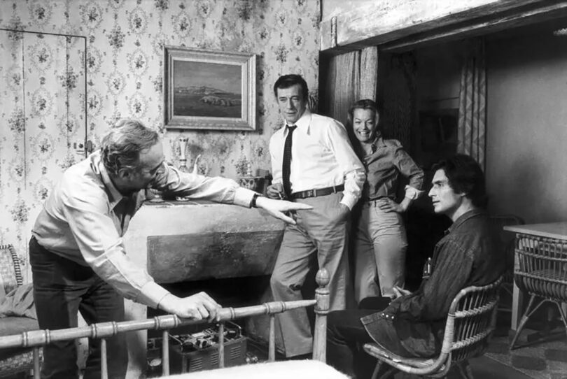 Black and white photo of four adults in a vintage living room, one man poking a fire, another leaning against a door frame, and two others observing him.
