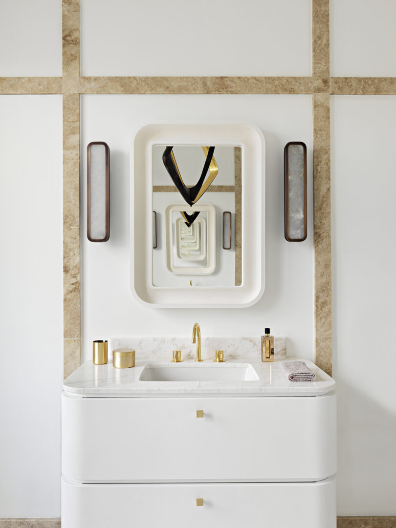 A modern bathroom vanity with a white sink, gold fixtures, and a square mirror framed by stone tiles. two vertical sconce lights flank the mirror.
