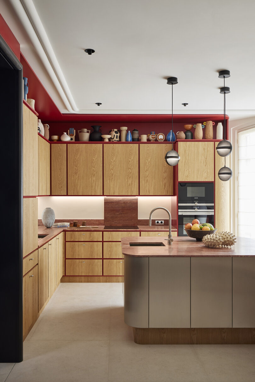 Modern kitchen with wooden cabinets, red accents, and an island under pendant lights, featuring built-in appliances and decorative items.