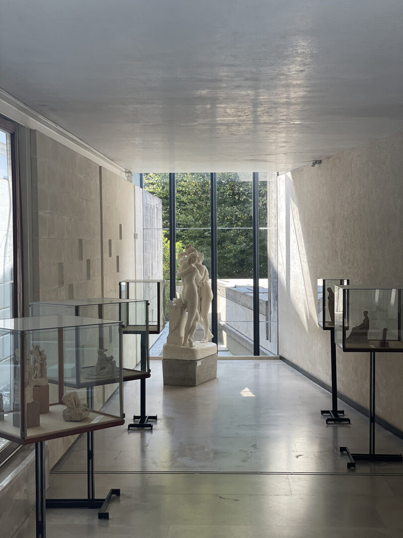A sunlit art gallery with a white marble statue in the center, surrounded by glass display cases with sculptures.
