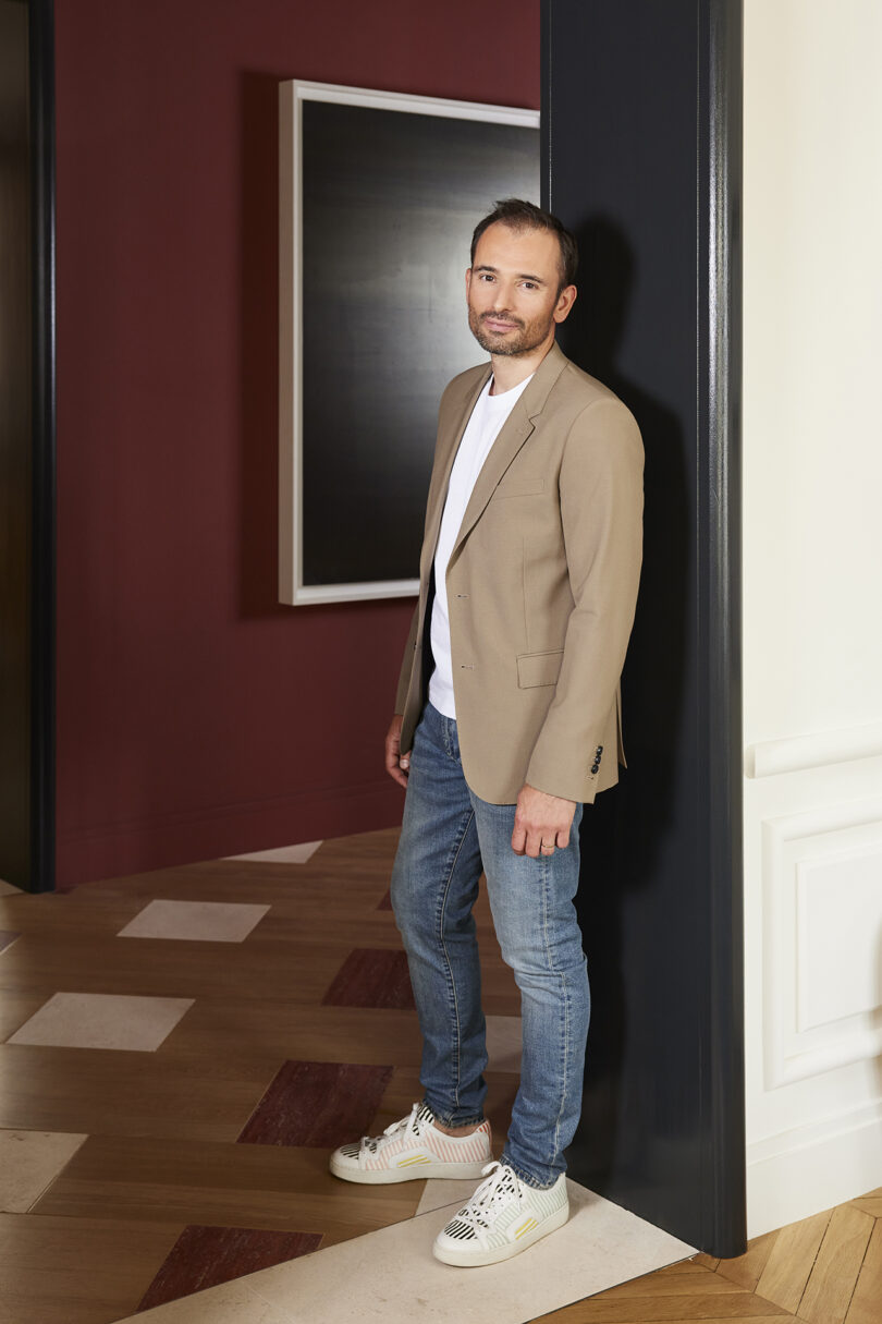 A man in a beige blazer and jeans stands in a doorway with a dark red wall and parquet flooring behind him.