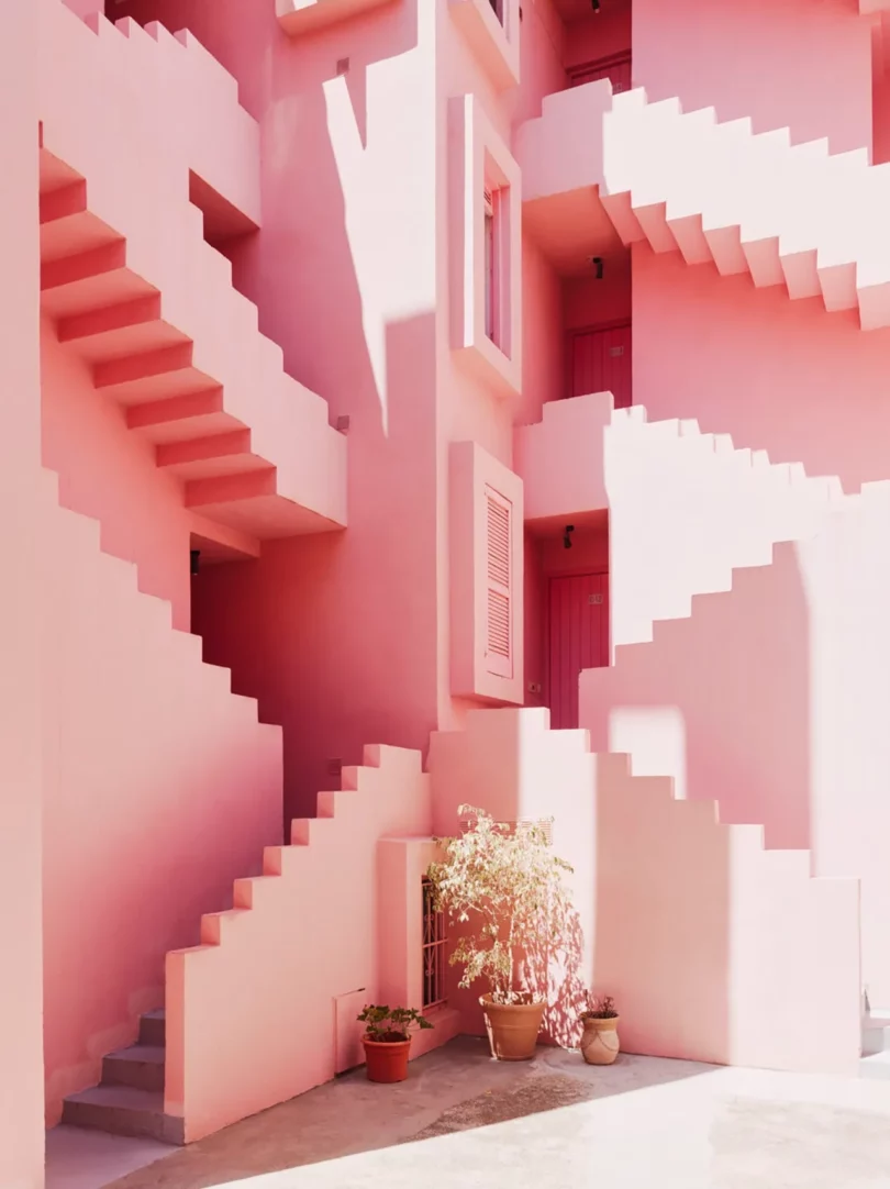 Pink architectural structure featuring staggered staircases and potted plants, highlighted by soft shadows and sunlight.