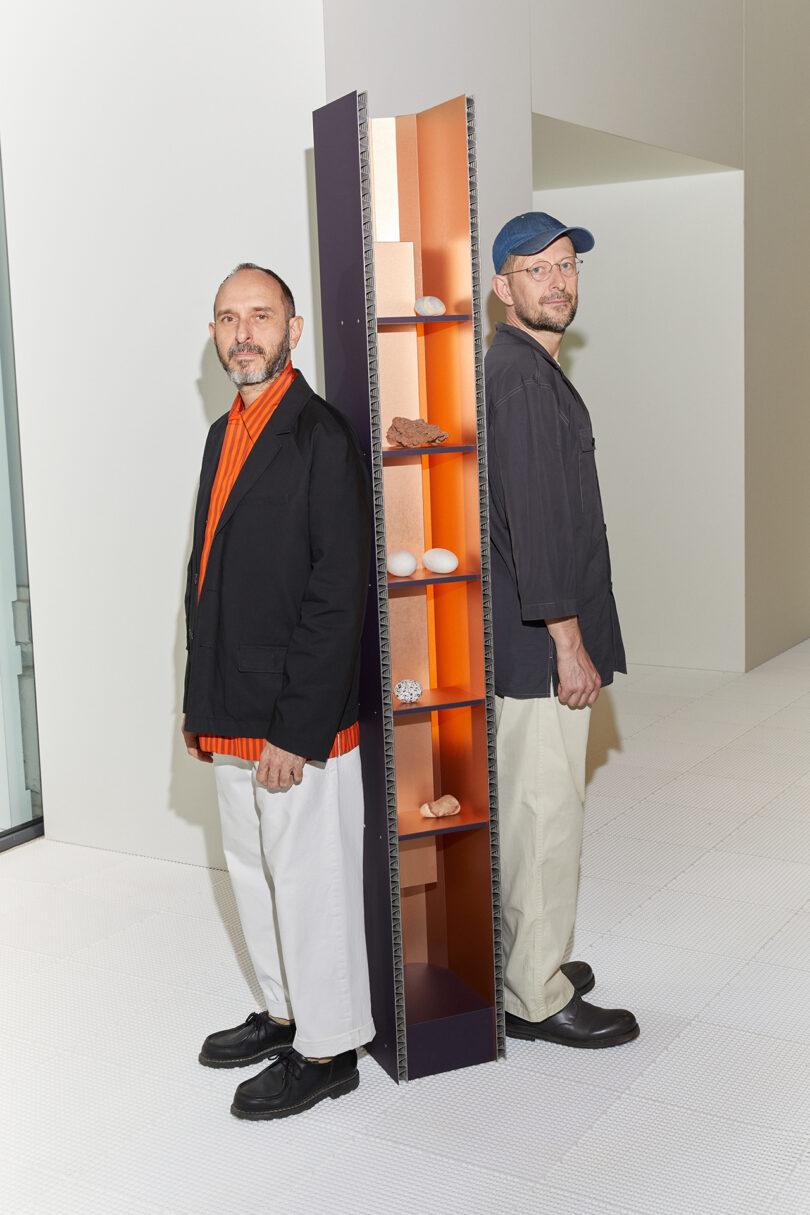 Two men standing back-to-back, one facing forward and one backward, by a colorful slim shelving unit filled with shoes.