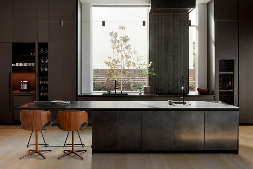 Modern kitchen with dark cabinetry and a central island featuring bar stools and a view of an outdoor garden through a large window.