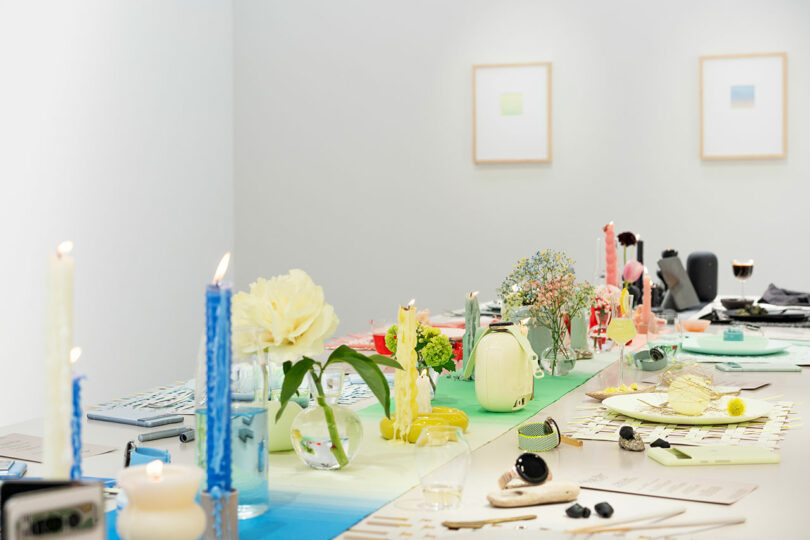 A modern dining table setup displaying an eclectic mix of colorful vases, flowers, candles, and place settings in a bright room with minimalist wall art.