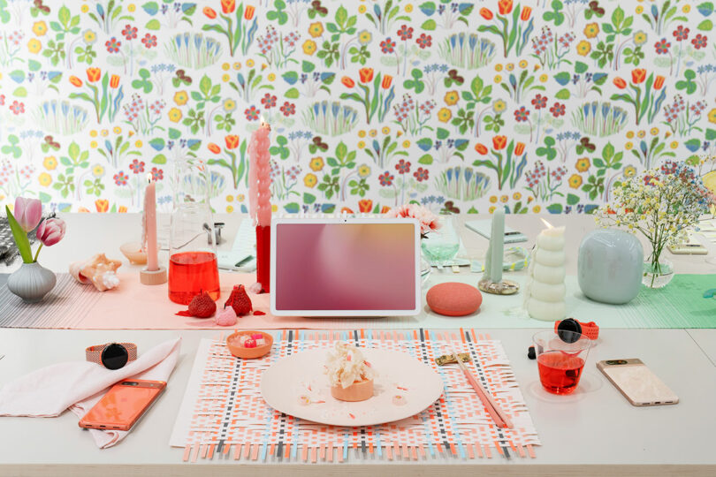 A colorful dining table setup featuring a Google laptop, wildflowers, and a partially eaten cake, with vibrant floral wallpaper in the background.