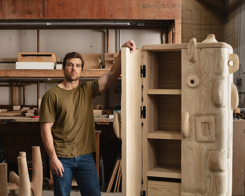 A man standing next to a wooden cabinet in a woodworking workshop, looking at the camera.