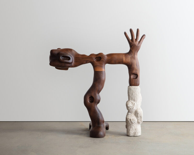 A wooden sculpture of abstract, human-like figures intertwined, featuring detailed hands and facial features, against a blank background.
