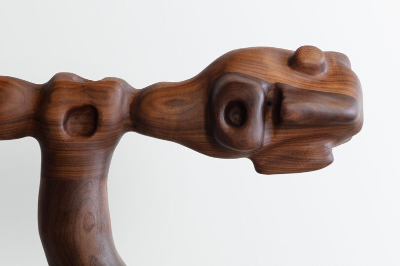 Detail of a wooden sculpture of abstract, human-like figures intertwined, featuring detailed hands and facial features, against a blank background.