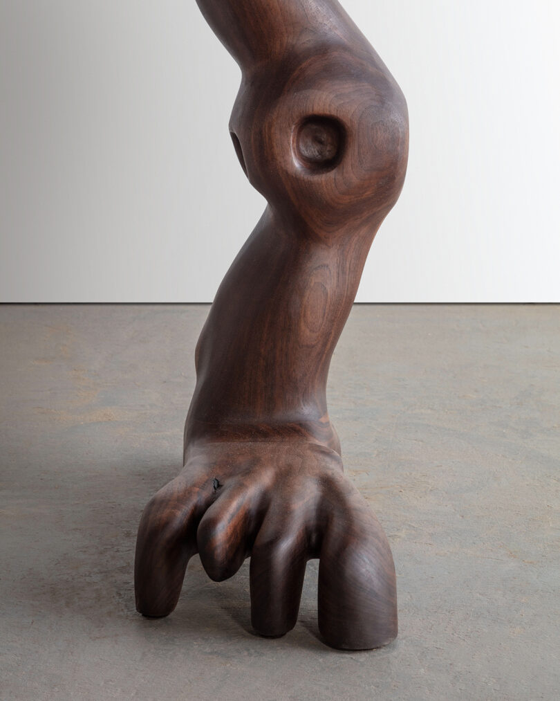 Detail of a wooden sculpture of abstract, human-like figures intertwined, featuring detailed hands and facial features, against a blank background.