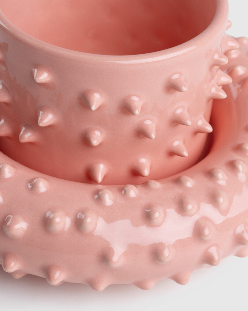 A pink ceramic Gustaf Westman mug and saucer with a textured, spiked surface stacked atop each other against a white background.
