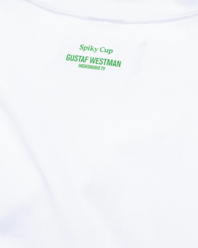 Close-up of a white t-shirt with green text "spiky cup Gustaf Westman highsnobiety" embroidered on the chest.