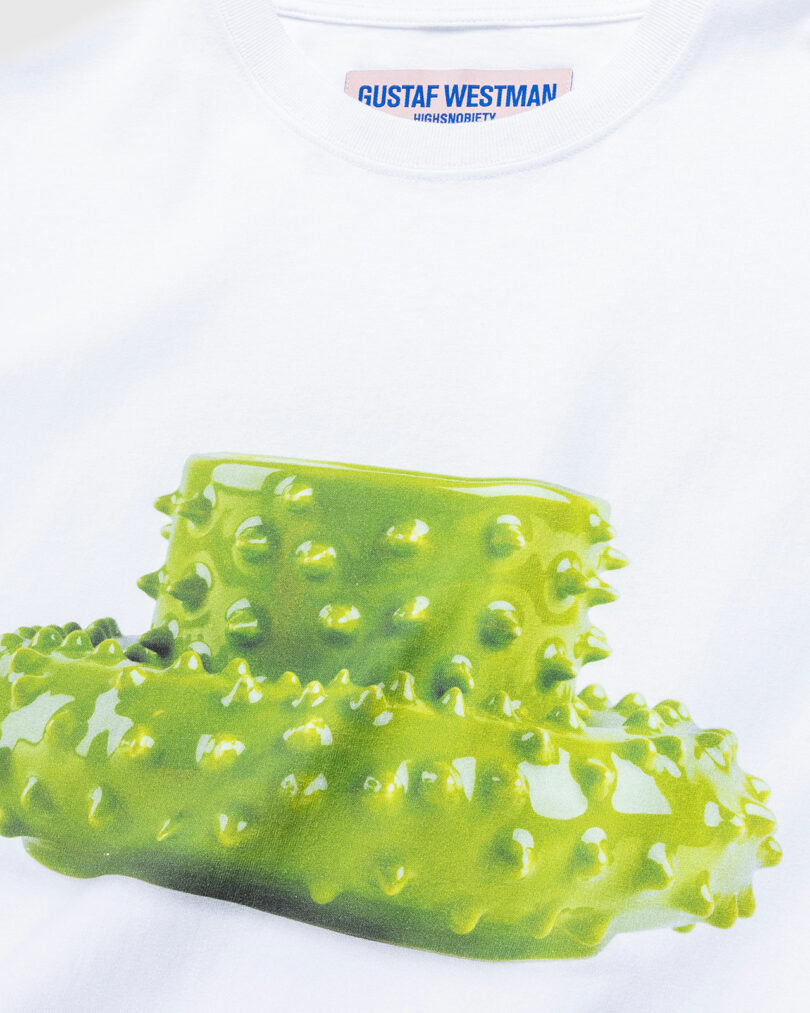 A white t-shirt featuring a graphic print of a vibrant green Gustaf Westman mug and saucer with a glossy texture. The tag reads "Gustaf Westman Highsnobiety."