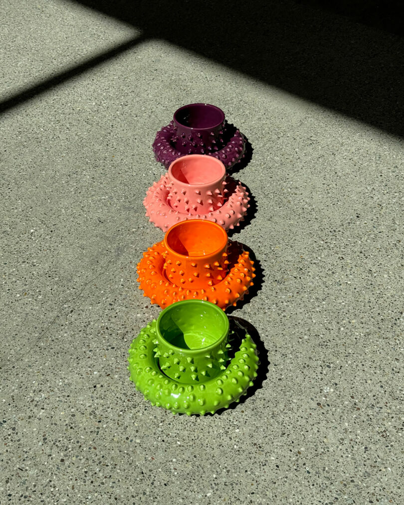 Five colorful, spiky Gustaf Westman mugs and saucers in shades of green, orange, pink, purple, and red, arranged in a line on a sunny