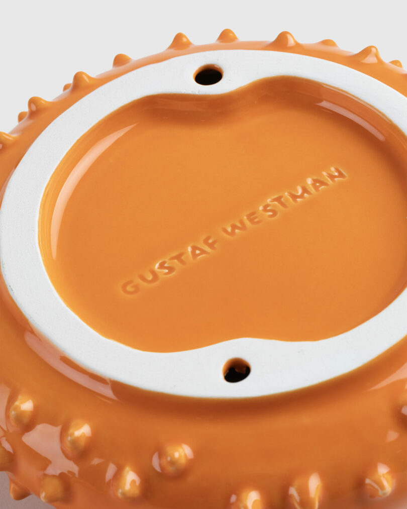 Close-up of an orange ceramic mug and saucer designed by Gustaf Westman with the name "Gustaf Westman" embossed in the center, featuring a spiked edge design.
