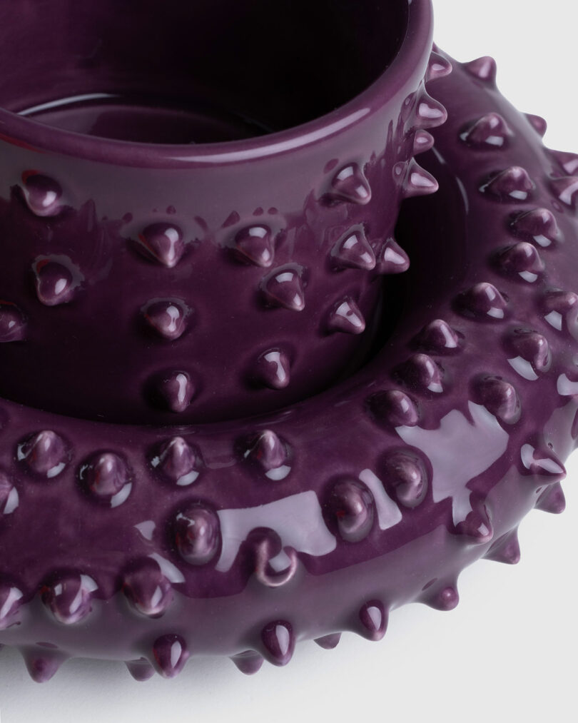 A purple ceramic Gustaf Westman mug and saucer with a textured, spiked surface stacked atop each other against a white background.