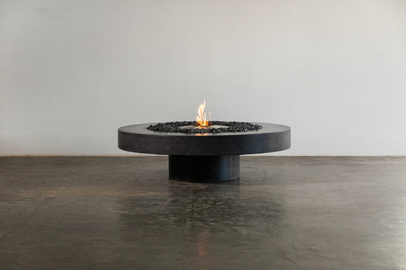 A modern low-standing James de Wulf fire pit table with a small flame in the center, set against a plain background.