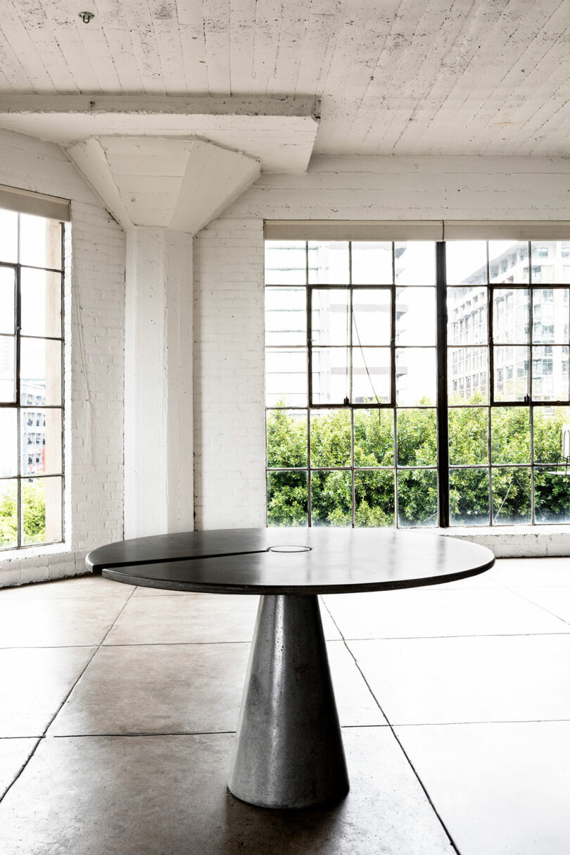A minimalist room with white walls, large windows, and a round black James de Wulf table.