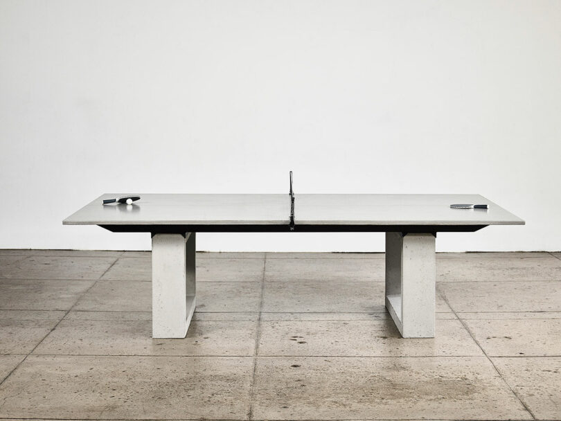 Minimalist concrete ping-pong table with two paddles and a ball on an industrial floor.