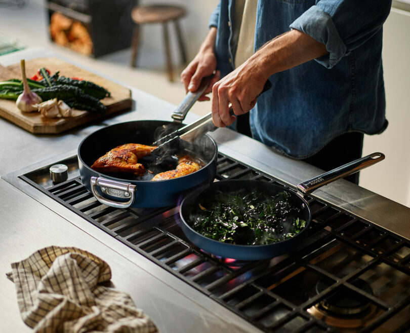A person sautéing chicken in a pan next to another pan with greens on a stovetop.