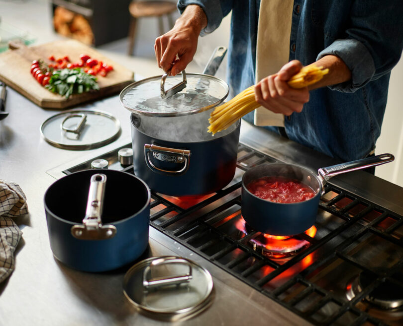 A person is cooking pasta over a stove, with a pot of boiling water and a separate saucepan with tomato sauce.