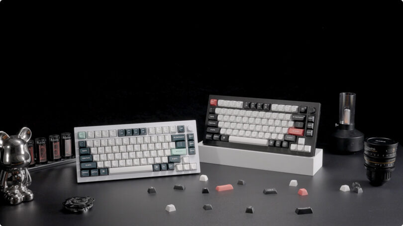 Two Keychron Q1 HE mechanical keyboards on a desk surrounded by loose keycaps, camera lenses, and a silver teapot, against a black background.