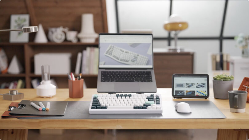 Keychron Q1 HE mechanical keyboard set in front of a MacBook on stand on a desk with an iPad and white mouse to the right and pens, files, and pen holder to the left.