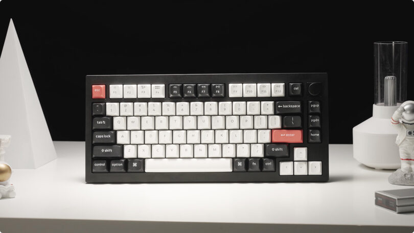 A Keychron Q1 HE mechanical keyboard with white and red keys on a desk flanked by a small white statue and a vase.
