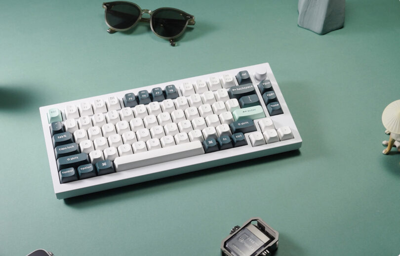 A stylish Keychron Q1 HE mechanical keyboard with white and teal keys on a green desk surrounded by sunglasses, a camera and a small chair.