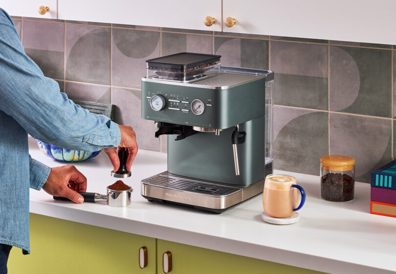 Person using a KitchenAid Espresso Collection machine to make coffee in a colorful kitchen, with a cup of coffee and coffee beans nearby.