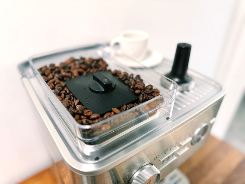 A KitchenAid Espresso Collection coffee grinder filled with beans, featuring a transparent lid, with a tamper, white cup and saucer in the background.