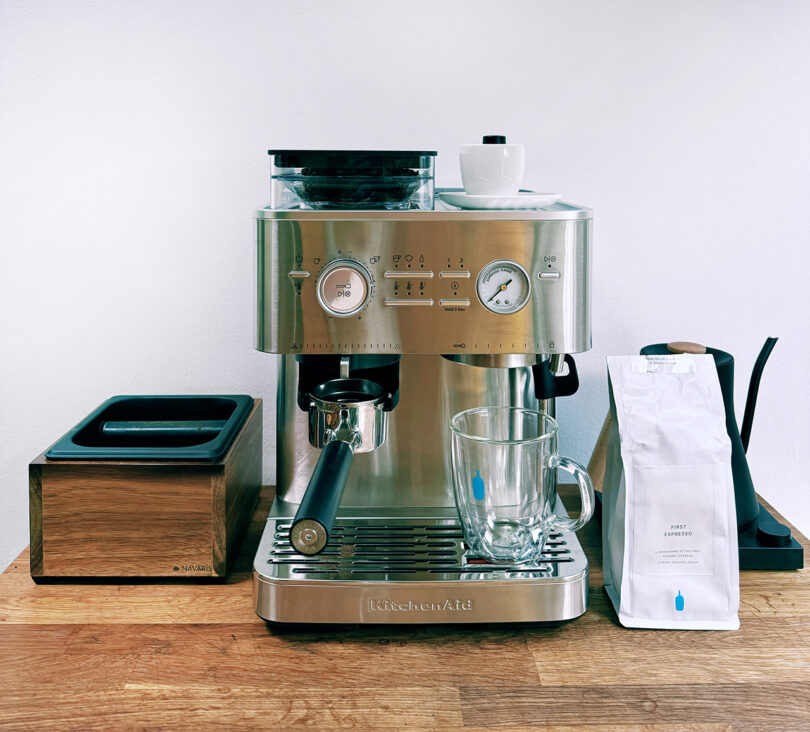 A close-up of a KitchenAid Espresso Collection machine with a bag of Blue Bottle espresso beans, knock box, and Fellow Kettle on each side.