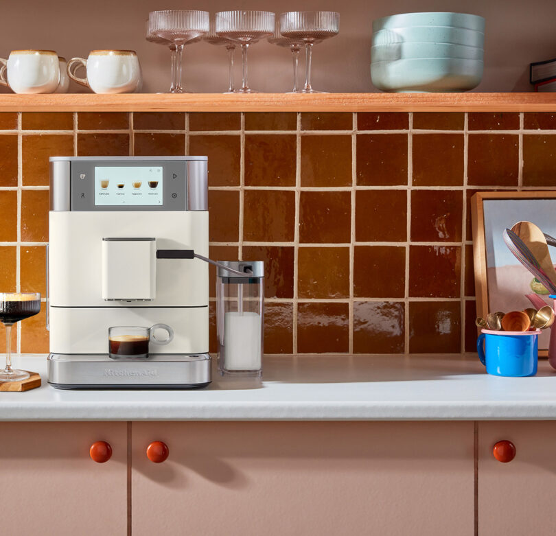 A modern KitchenAid Espresso Collection coffee machine on a kitchen counter brewing a cup of coffee, with an attached milk container on the side with decorative items nearby.