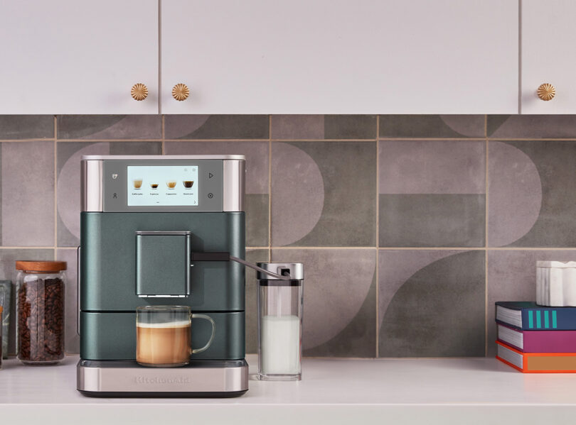 A modern KitchenAid Espresso Collection coffee machine on a kitchen counter brewing a cup of coffee, with a container of milk on the side and decorative items nearby.