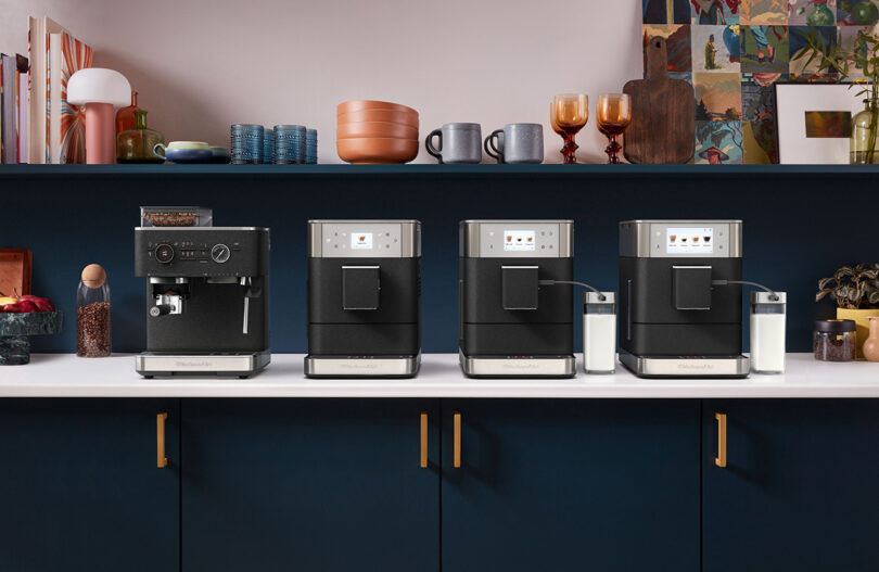 Four modern KitchenAid Espresso Collection coffee machines on a dark kitchen counter, flanked by cabinets and decorative items.