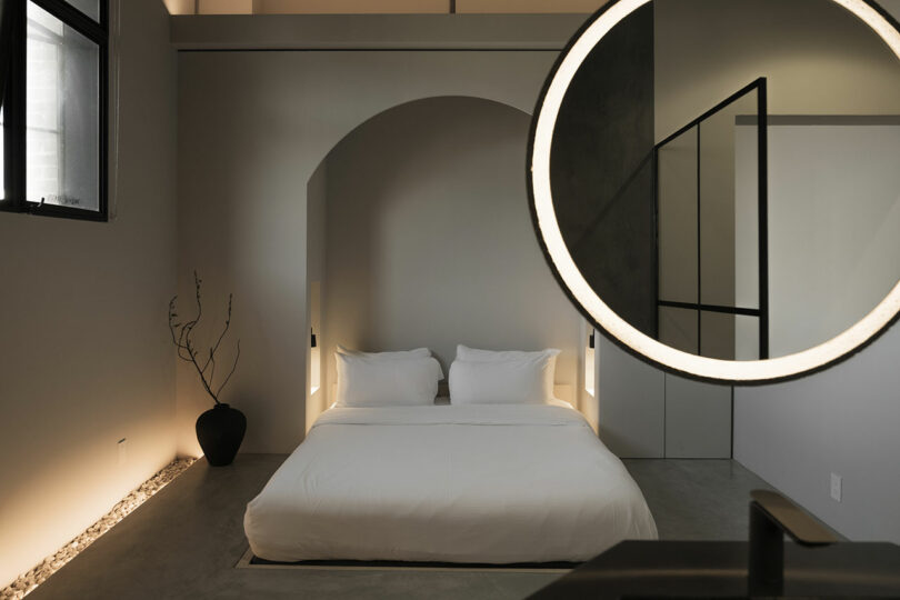 Modern bedroom with minimalistic design featuring an arched doorway, large circular mirror, and soft lighting.