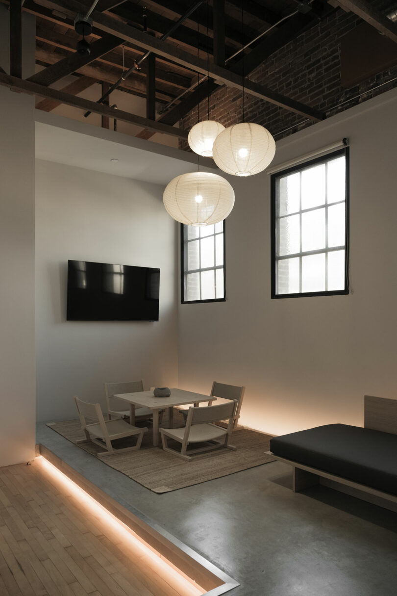 Minimalist living room with paper pendant lamps, a black flat-screen TV, and simple furniture against exposed brick.