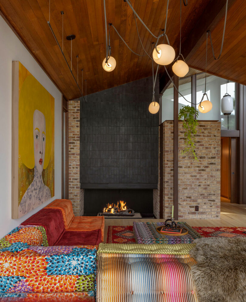 Modern living room with a colorful patchwork rug, red bed, and a fireplace, complemented by a large portrait painting on the wall.