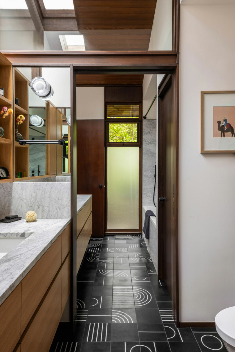 Narrow modern bathroom with black and white patterned floor, wooden cabinetry, and frosted glass door leading to another room. natural light streams in through skylights.