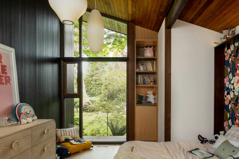 Cozy bedroom with large windows showing a garden view, wooden interiors, and colorful wall decorations.