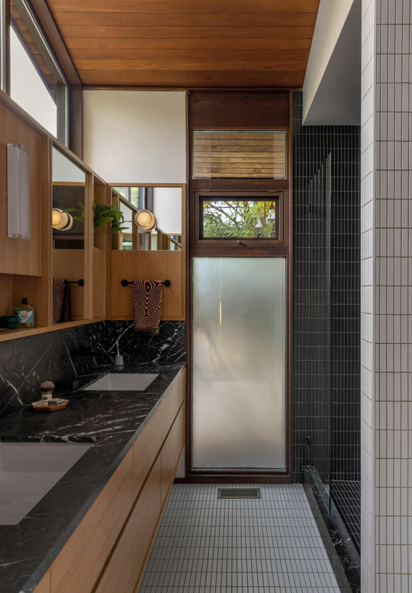 Modern bathroom with black marble countertop, wooden cabinets, frosted glass door shower, and white tiled wall.