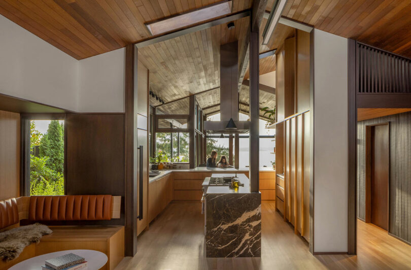 Modern kitchen with wooden ceilings and floors featuring a marble island, surrounded by open plan living space with natural light and greenery visible outside.
