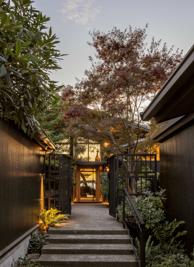 A serene pathway between black wooden fences leading towards a warmly lit house, flanked by lush greenery and trees under a dusky sky.