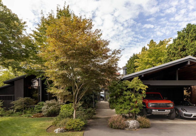 Two modern black houses with a connecting walkway, surrounded by lush trees and a driveway with two cars, one red and one silver.