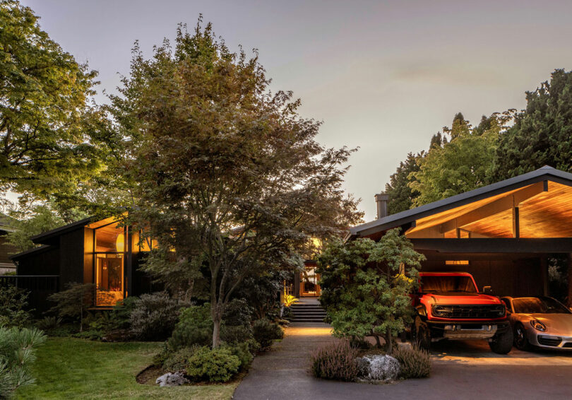 Modern suburban home with a lush garden during sunset, featuring a carport with two cars parked under a well-lit overhang.